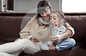 Young mother with her daughter are reading a book at home while sitting on the couch