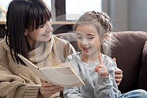 Young mother with her daughter are reading a book at home while sitting on the couch