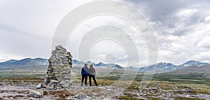 Mom and daughter stands on a mountain top next to a cairn with looking at beautiful mountain view of Rondane national park