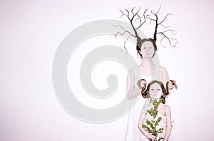 Mom and daughter in white dresses on a white background depict winter and spring, holding flowers and a twig with leaves.