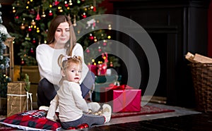 Mom and daughter in white sweaters sitting on the carpet near the Christmas tree. gifts, lights, red balls in the background