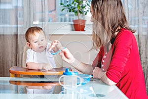 Mom feeding the baby holding hand with a spoon of food in the kitchen. Healthy baby nutrition. Emotions of a child