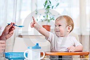 Mom feeding the baby holding hand with a spoon of porridge in the kitchen. Emotions of a child while eating healthy food