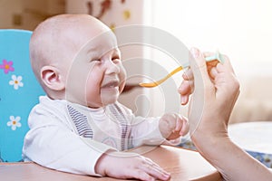 Mom feeds by spoon an emotional infant boy with blue eyes at day time