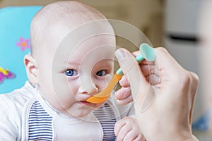 Mom feeds by spoon an emotional infant boy with blue eyes at day time