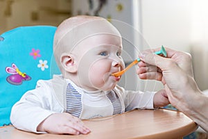 Mom feeds by spoon an emotional infant boy with blue eyes at day time