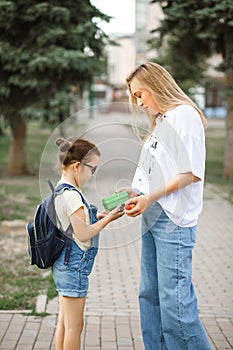 Mom gives daughter cute little schoolgirl with backpack lunch box. Near by school outdoor. Food for kids