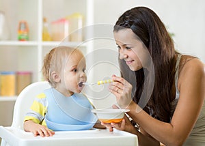 Mom giving homogenized food to her baby son on high chair in kitchen.