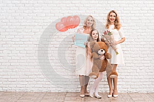 Mom, grandmother and girl are posing together near a light wall.