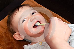 Mom hand is brushing the teeth of the happy toddler baby boy. The mother conducts oral hygiene of the smiling child lying on a