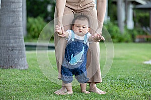 Mom helping and support baby learn to walk with love outdoors on grass. First steps of baby learning walk with family. Adorable