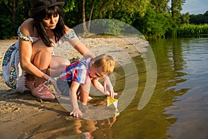 Mom helps the little boy to lower the paper boat to the water