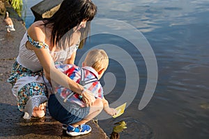 Mom helps the little boy to lower the paper boat to the water.