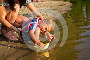 Mom helps the little boy to lower the paper boat to the water