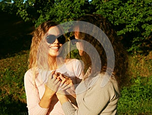 Mom and her adult daughter hold hands in a summer park, happy women wearing sunglasses