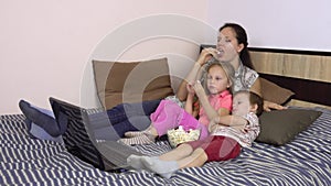 Mom and her children are watching funny cartoons on a laptop in bed and eating popcorn together.