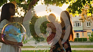 Mom and her daughters blow big soap bubbles in evening city park at sunset. Children and mother play together and laugh