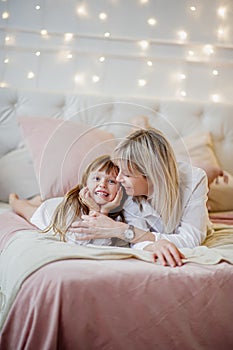 Mom and her little daughter have fun and play sitting on the bed in a bright bedroom with Christmas decor
