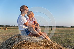 Mom and her little son are sitting in a field with hay rolls in the setting sun. A woman in denim shorts and a white t-shirt.