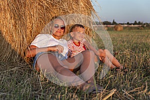 Mom and her little son are sitting in a field with hay rolls in the setting sun. A woman in denim shorts and a white t-shirt.