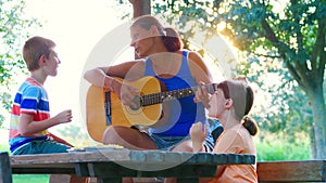 Mom and her two children sing songs together playing guitar outdoors on a sunny day.