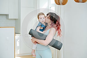 Mom holding her baby girl, kissing her, holding a yoga mat in the kitchen