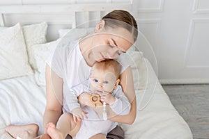 Mom holds baby in her arms with a rodent on a white bed with cotton bedding at home, teether