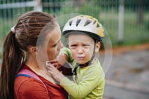 Mom holds a crying todler in her arms. The boy has a helmet on his head. They are on the street.