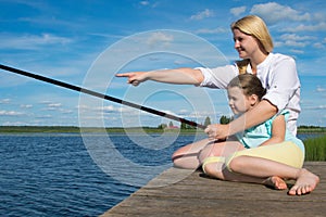 Mom holds the fishing rod and hand shows her daughter on the catch, on the pier, against the backdrop of a beautiful landscape,