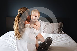 Mom hugs nine-month-old baby girl sitting with her back to camera. Mother with child play together at home sitting on bed