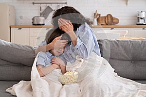 Mom and kid son with bowl of popcorn watching scary movie closing their eyes sitting on sofa at home