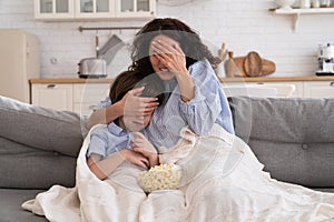 Mom and kid son with bowl of popcorn watching scary movie closing their eyes sitting on sofa at home