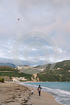 Mom with a kite soaring in the sky walks along the beach with a little girl. Back view