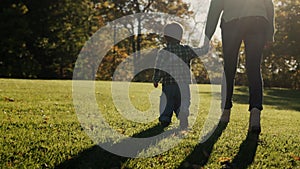 Mom leads the baby by the hand, walking next to the green grass in the rays of the setting sun. Follow shot