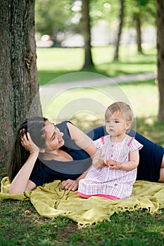 Mom lies next to a little girl sitting on a blanket on the green grass