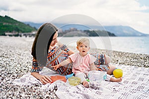 Mom lies next to a sitting little girl on a blanket on a pebble beach