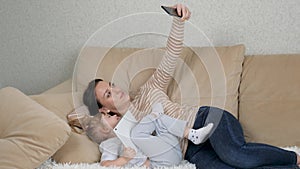 Mom with a little child take a selfie on a smartphone while lying on the couch and laugh, family photo sessions at home