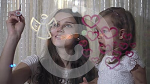 Mom with little daughter draw heart and angel paints on glass.