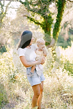 Mom with a little daughter in her arms stands near a tree twined with ivy