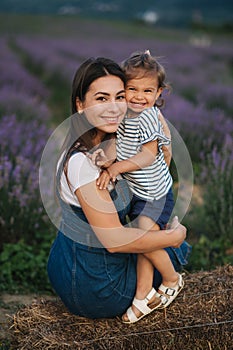 Mom and little daughter sits on hay by the farm. Background of summer lavender field. Family denim style. Little girl