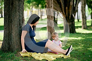 Mom with a little girl are sitting on a blanket on a green lawn under a tree