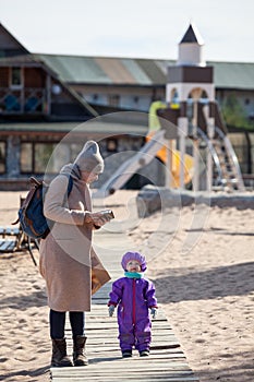 Mom looks at a three-year-old child dressed in warm, windproof overalls standing on wooden walkways. Walk on the autumn beach