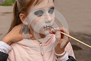 Mom painting daughter face for Halloween party in the courtyard of his house. Children`s Zombie Makeup. Halloween and day of the