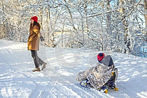 Mom is pulling a child on a sledge walking on a winter Sunny day out of doors. Baby wrapped in a blanket rides on a sled