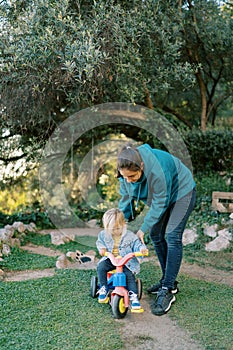 Mom pushes a little girl pedaling a bicycle on the lawn in the park