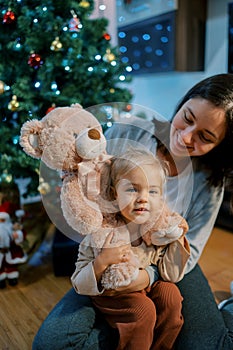 Mom put a teddy bear on the neck of a little girl sitting on the floor near the Christmas tree