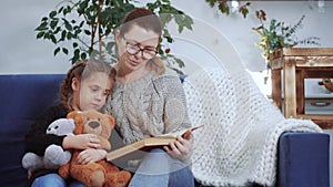 Mom read a book to her daughter at home. Kid dream coronavirus stay home concept. Mom is sitting on the couch with