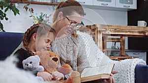 mom reads a book to her daughter at home. lockdown kid dream coronavirus stay home concept. mom is sitting on the couch