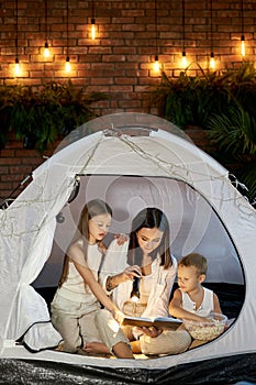 Mom reads children a bedtime story sitting in a tent at home. Mother son and daughter hug and read a book with a flashlight