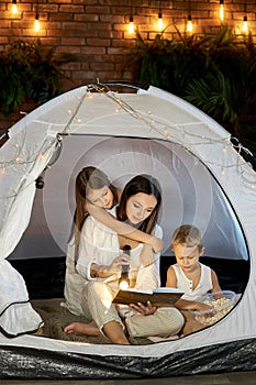 Mom reads children a bedtime story sitting in a tent at home. Mother son and daughter hug and read a book with a flashlight